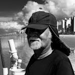 Black and white portrait of an older fisherman with a beard on Praia Grande, São Paulo. Urban coast and cloudy sky in background.